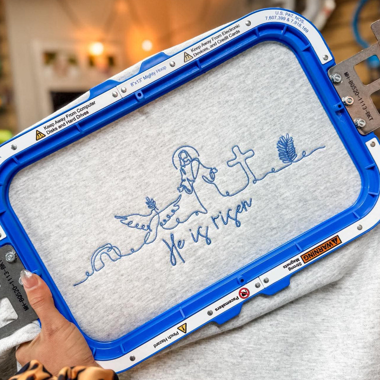 Embroidery hoop with a design on a gray fabric held by a person in a workshop setting.