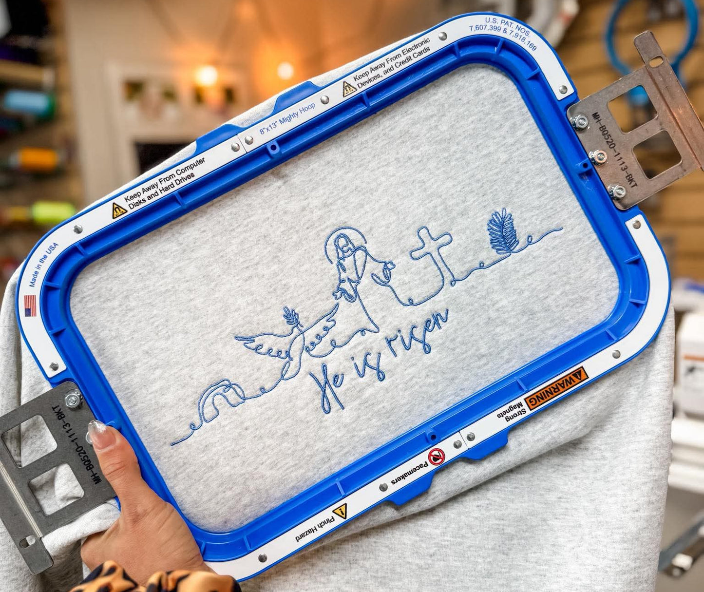 Embroidery hoop with a design on a gray fabric held by a person in a workshop setting.