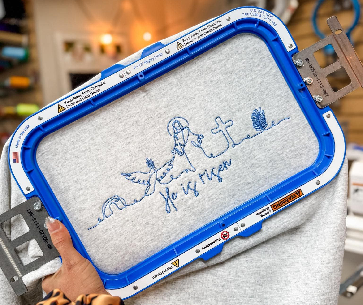 Embroidery hoop with a design on a gray fabric held by a person in a workshop setting.